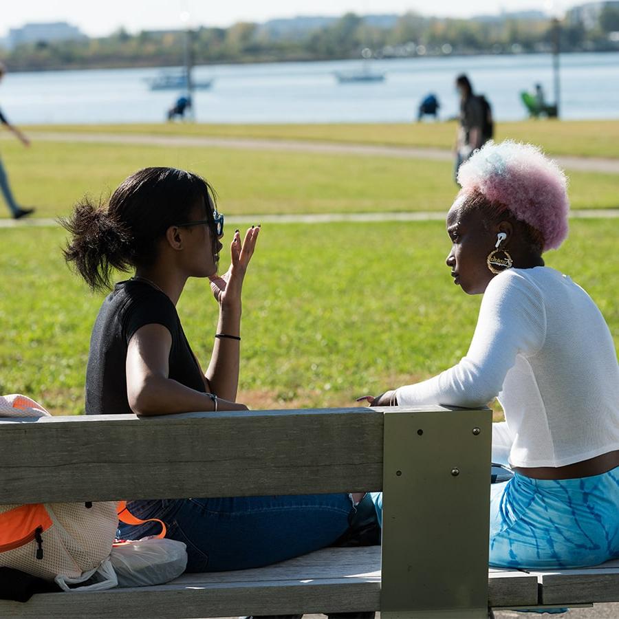 Two girls talk on bench campus lawn